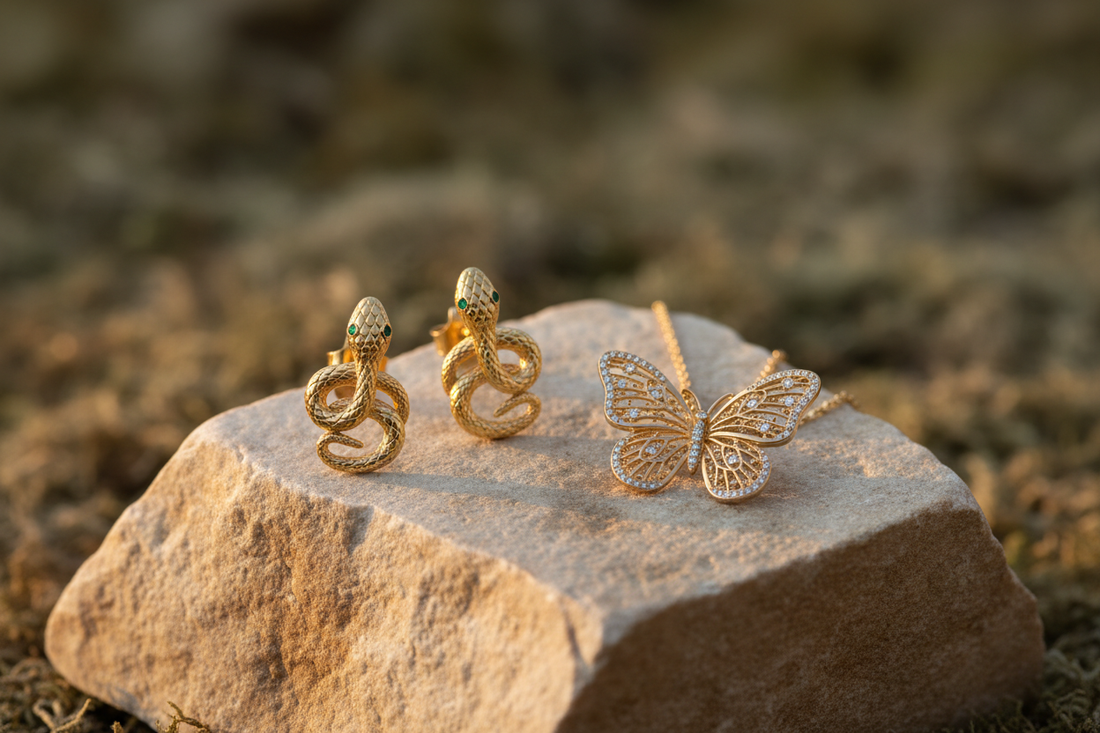 Bodegón de joyería mostrando pendientes de serpiente dorados y un colgante de mariposa sobre una piedra natural